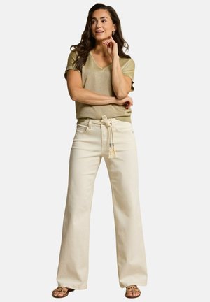 Vrouw met lang donker haar, gekleed in een beige top met korte mouwen, creme wijde broek met hoge taille en een koordriem, en sandalen.