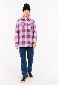 Moteris, vilkinti rožinės ir purpura dėmėtos mygtukinės striukės, tamsiai mėlynų džinsų, juodo kepurės ir juodų batų su smėlio spalvos padais, stovi ant baltos fono.