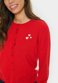 Gai cardigan rouge avec un tricot texturé, comportant une rangée de boutons et trois cœurs blancs brodés sur le côté gauche.