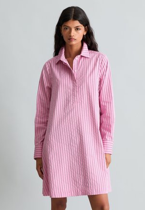 Femme portant une robe-chemise rose à manches longues avec de fines rayures verticales noires, debout devant un fond gris clair uni.
