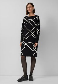 Czarna dzianinowa sukienka z długimi rękawami, zdobiona białymi abstrakcyjnymi wzorami. Stylizowana z nieprzezroczystymi czarnymi rajstopami i czarnymi loafersami. Minimalistyczny design.