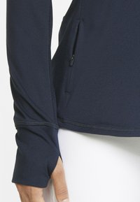 Maglia a maniche lunghe blu navy con un design fitted, dotata di una tasca laterale con zip e aperture per il pollice nei polsini per una maggiore funzionalità.