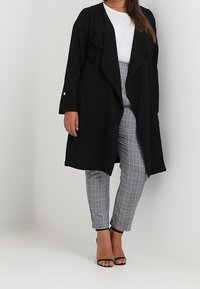 Černý dlouhý kabát s volnou přední částí, kombinovaný se šedými kostkovanými kalhotami. Oblečení zahrnuje jednoduchý bílý top a černé sandály na podpatku.