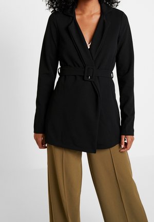 Frau trägt einen schwarzen Blazer mit Gürtel und Revers sowie beige weite Hosen vor einfarbigem Hintergrund.