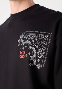 Sweat-shirt noir avec motif paisley et toile d'araignée blanc et texte rouge "HUGO" sur la partie supérieure de la poitrine, près de l'encolure.
