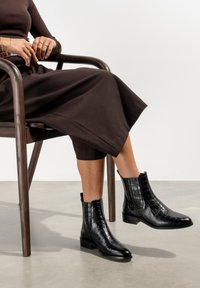 Schwarze Lederstiefeletten mit Krokodilstruktur, quadratischer Zehenpartie und niedrigem Blockabsatz, kombiniert mit einem braunen Rock, sitzend auf einem Holzstuhl.