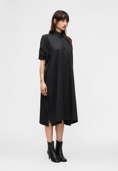 Γυναίκα με μακριά σκούρα μαλλιά, φορούσα μια χαλαρή μαύρη πουκαμίσ α-φόρεμα με κουμπιά και μαύρα μποτάκια αστραγάλου, στέκεται μπροστά σε απλό φωτεινό φόντο.