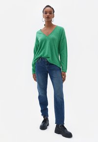 Maglione verde oversize a V in maglia texturizzata, abbinato a jeans blu a gamba dritta e stivali neri con lacci.