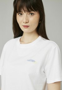 T-shirt en coton blanc avec un design graphique de texte bleu et jaune sur le côté gauche de la poitrine. Manches courtes et col rond.