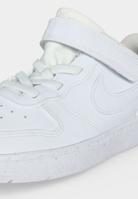 Hvid Nike-sneaker med snørebånd og en velcrolukning, der viser detaljerede syninger og perforeringer på tåområdet.