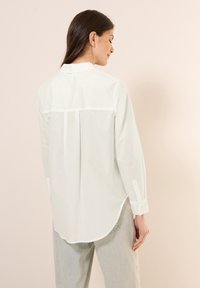 Camicia bianca con bottoni, realizzata in tessuto liscio, con orlo curvo, fessure laterali e yoke posteriore strutturato. Presenta maniche lunghe con polsini.