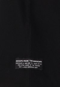Zwarte stof met een gladde textuur en een rechthoekig label. Op het label staat witte tekst met de woorden "ESCAPE FROM / THE UNKNOWN."