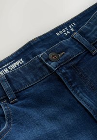 Džínsy v tmavomodrom odtieni s boot fit strihom, so zapínaním na gombík, pútkami na opasok a kontrastným prešívaním pozdĺž vreciek a švov.