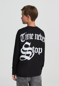 Czarna koszulka z długim rękawem z dużym białym napisem na plecach "Czas nigdy nie stoi" w postarzałej czcionce. Prosty, casualowy design.