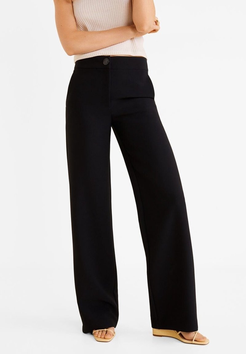 Un pantalon noir à jambes larges en tissu lisse, avec une taille haute et une fermeture à un bouton à l'avant. Associé à des sandales légères.