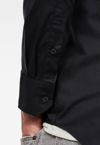 Camicia nera a maniche lunghe con polsini con bottone, realizzata in materiale lucido. Le caratteristiche principali includono una cucitura diagonale e una vestibilità su misura. Jeans grigi visibili in vita.