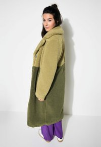 Cappotto in shearling bi-tono verde e oliva con colletto a scollo a V, grandi bottoni e tasche laterali; modello che indossa pantaloni viola e sneakers.