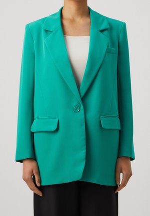 Femme portant un blazer bleu sarcelle vif sur un haut blanc, avec un pantalon noir, debout devant un fond clair uni.