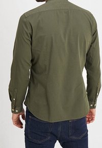 Mand i en olivengrøn langærmet skjorte og mørkeblå jeans, vist fra ryggen mod en ensfarvet lys baggrund.