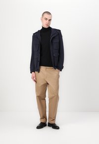 Marineblauwe multi-pocket jacket over een zwarte coltrui, gecombineerd met tan broek en zwarte enkellaarzen. Glad materiaal met een relaxte pasvorm.