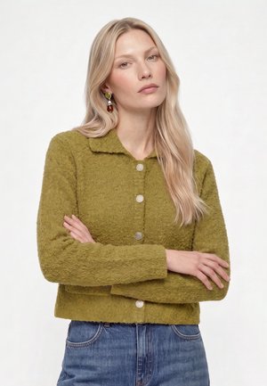 Lyshåret kvinde med langt hår iført grøn tekstureret jakke med knapper og blå jeans, armene krydset, neutralt ansigtsudtryk, hvid baggrund.