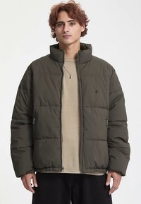 Olive green puffer jacket with a zip front, high collar, and side pockets. Textured fabric with quilted pattern. Worn over a beige top.