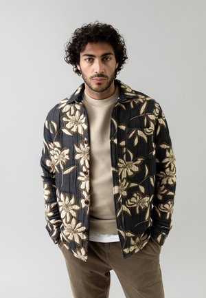 Floral-patterned jacket in black with beige flowers, featuring button closure and side pockets, layered over a beige shirt and brown pants.