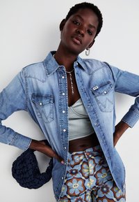 Camisa de ganga azul clara com bolsos, frente de botão e detalhes de costura. Usada sobre uma camisola cinza e combinada com calças de padrão floral.