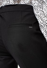 Svarta byxor med en slät tygtextur, utrustad med en bakficka och subtil varumärkesetikett. Tillsammans med en vit topp med svarta mönster.