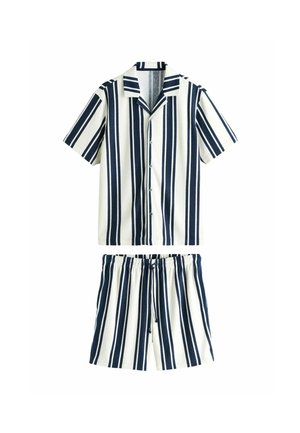 Ung mand, der står indendørs iført en skjorte med lodrette navy og hvide striber og matchende shorts med navy tøfler.