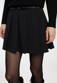 Gonna plissettata nera a righe verticali bianche sottili, indossata con collant neri e una cintura nera sottile, mostrata dalla vita fino a metà coscia.