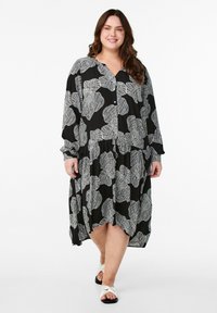 Schwarzes Kleid mit weißem Blattmuster, Knopfleiste, langen Ärmeln und plissiertem Rock. Kombiniert mit weißen Sandalen.