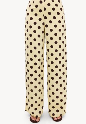 Pantalon beige à jambes larges avec de grands pois noirs, porté avec des sandales foncées à bout ouvert, visible de la taille jusqu'aux pieds.
