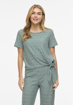 Žena s dugom plavom kosom, odjevena u svijetlozelenoj majici kratkih rukava i usklađenim hlačama s vezicom sa strane.