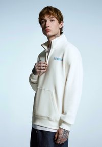 White zip-up pullover made of soft fabric, featuring a front pocket and blue logo on the chest. Model has tattoos and wears a gray bottom.