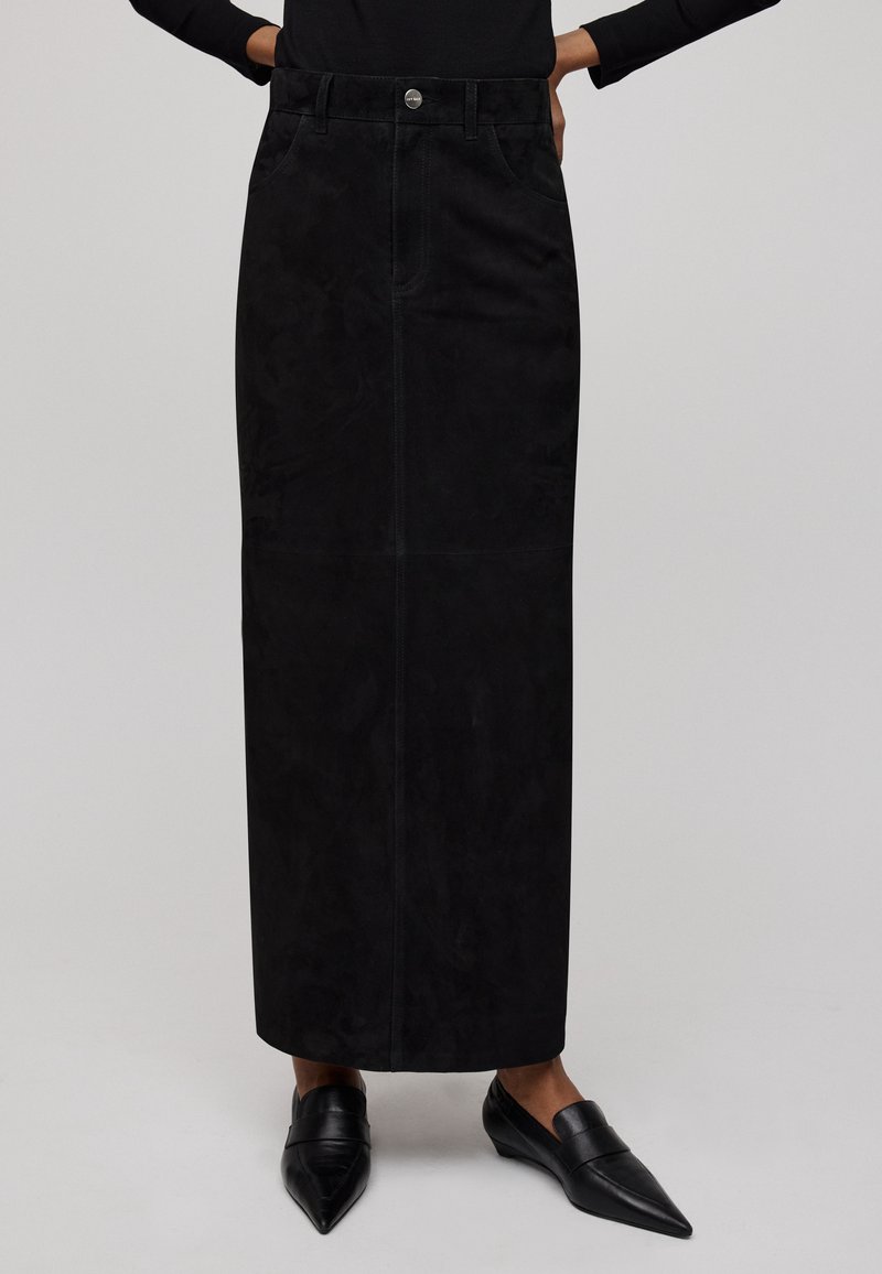 Schwarzer Wildleder-Maxxikleid mit geradem Schnitt, vorderen Taschen und einem einzigen Knopfverschluss. Getragen mit schwarzen spitzen Ballerinas.