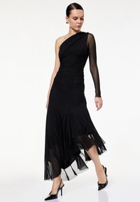 Sort kjole med én lang gennemsigtig ærme og rynk. Løstsiddende nederdel med lagdelt chiffon kant. Sammen med høje sorte sko.