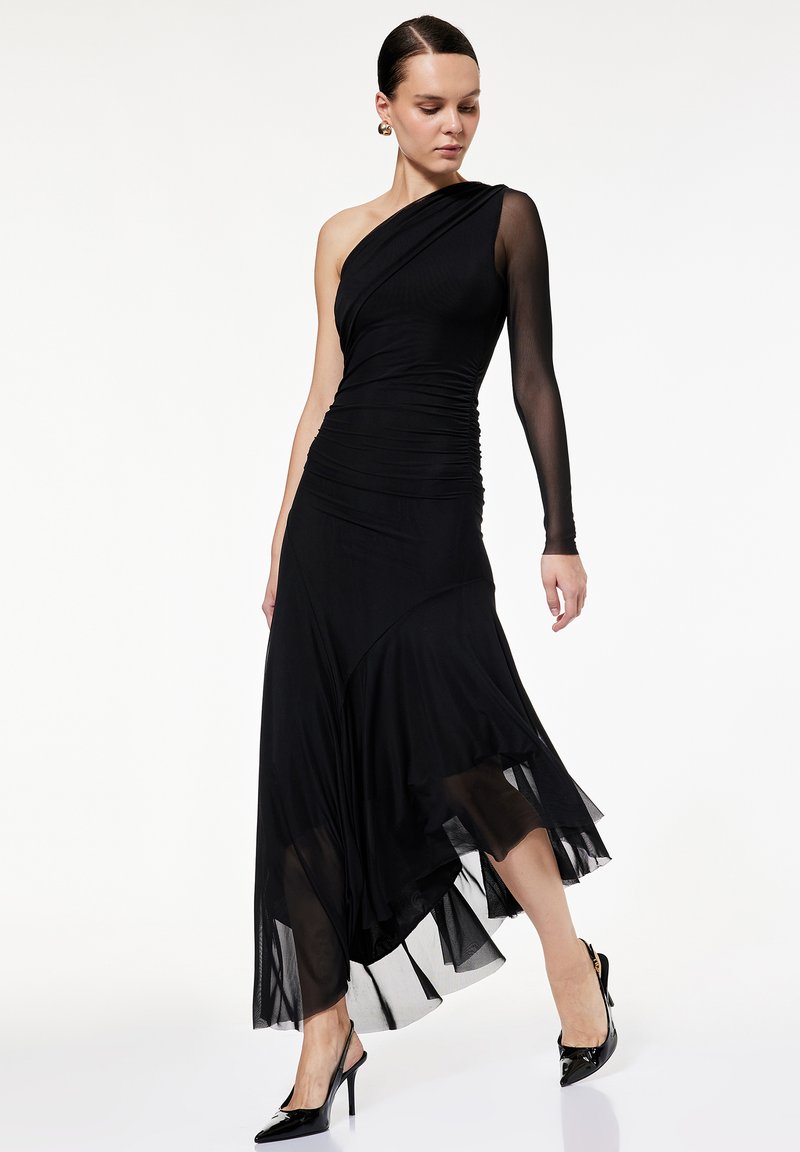 Sort kjole med én lang gennemsigtig ærme og rynk. Løstsiddende nederdel med lagdelt chiffon kant. Sammen med høje sorte sko.