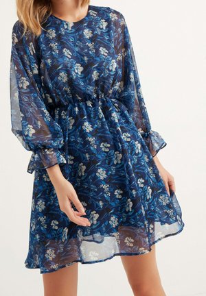 Vestido floral de tela navy transparente con flores azules y blancas. Presenta mangas largas y abullonadas, y una cinturilla elástica, fruncida en la cintura.