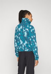 Fleecejacke in Türkis mit einem blauen abstrakten Muster, hohem Kragen und Logodetail auf dem Rücken. Sie hat eine lockere Passform und einen gerippten Saum.