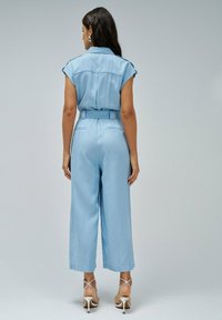 Combinaison en denim bleu clair avec des manches courtes, taille ceinturée, pantalon large et poches arrière. Texture lisse avec un design décontracté.