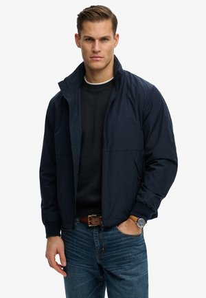 Mężczyzna noszący granatową kurtkę na zamek błyskawiczny na czarnym swetrze, niebieskie dżinsy, brązowy pasek i zegarek na nadgarstku, stojący z jedną ręką w kieszeni.