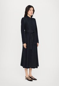 Mørke denim trenchcoat med dobbeltknaplukning, bælte i taljen og kontrasterende grøn syning. Modellen har sorte sko med stropper på.