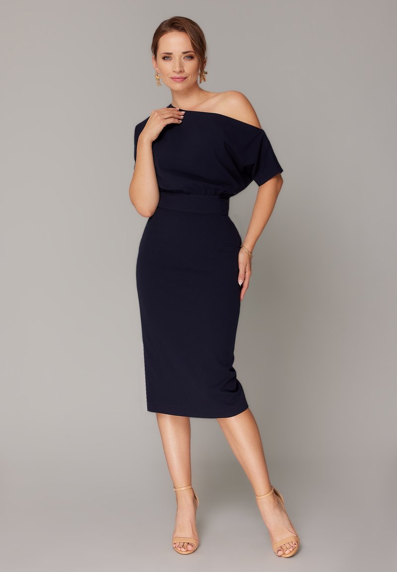 Granatowa sukienka bez ramiączek o dopasowanej sylwetce, z subtelnie zaznaczoną talią i długością do kolan. Gładki materiał o minimalnej teksturze, zestawiona z cielistymi szpilkami.