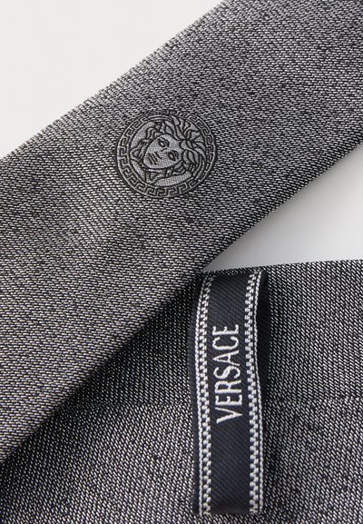 Siva tkanina z zapleteno teksturo, embosiranim logotipom Meduza in nalepko Versace. Oblikovanje poudarja mešanico razkošja in obrtniške izdelave.