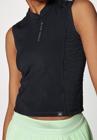 Gros plan d'une personne portant un haut sans manches Nike Trail noir avec des panneaux latéraux texturés et une taille de jupe plissée vert clair.