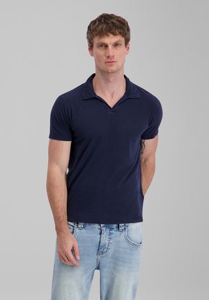 Junger Mann mit kurzen lockigen Haaren, der ein dunkelblaues Poloshirt mit kurzen Ärmeln und hellblaue Jeans vor einem einfarbigen grauen Hintergrund trägt.