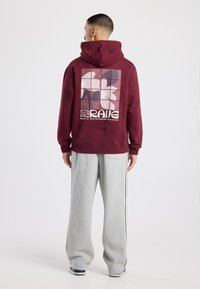 Sweat à capuche marron avec un imprimé graphique en rose et bleu dans le dos, présentant des formes géométriques et un texte audacieux. Associé à un pantalon de survêtement gris.