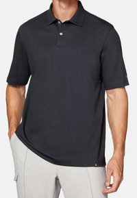 Polo noir avec col, manches courtes et texture subtile. Présente deux boutons blancs et une petite étiquette de marque au bas.