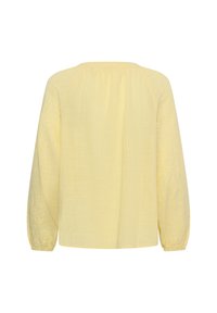 Blouse jaune clair à manches longues avec une texture florale subtile et des poignets élastiques, vue de dos sur fond blanc.
